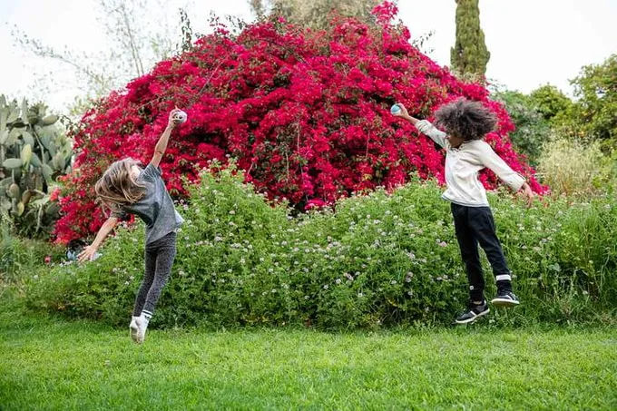 Two children throwing and catching a Storyball to each other on a lawn in front of a bank of red flowers